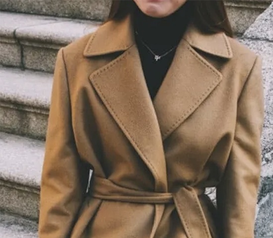 Woman wearing a camel-colored wool coat with wide lapels and belted waist, layered over a black turtleneck sweater on stone steps
