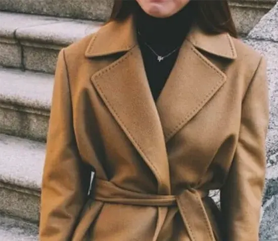 Woman wearing a camel-colored wool coat with wide lapels and belted waist, layered over a black turtleneck sweater on stone steps