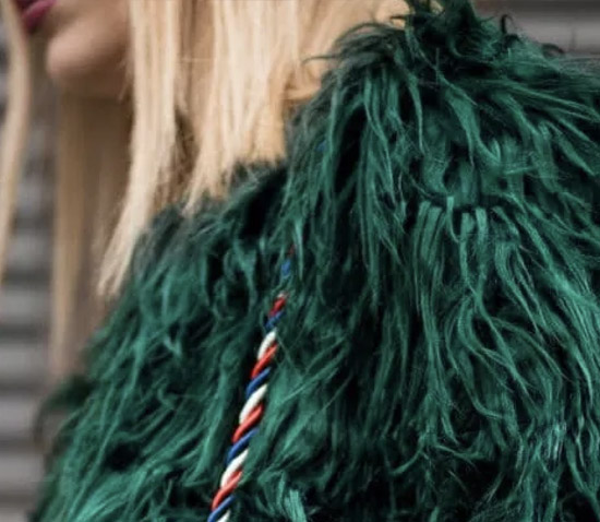 Woman wearing a bold green faux fur coat with long textured strands and a twisted red, white, and blue shoulder strap detail