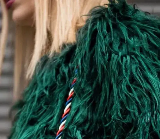 Woman wearing a bold green faux fur coat with long textured strands and a twisted red, white, and blue shoulder strap detail