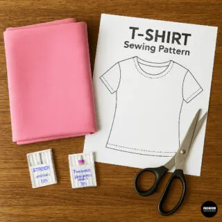 Flatlay of folded pink cotton jersey, printed T-shirt sewing pattern, two packs of stretch sewing machine needles, and sharp fabric scissors on a wooden table.