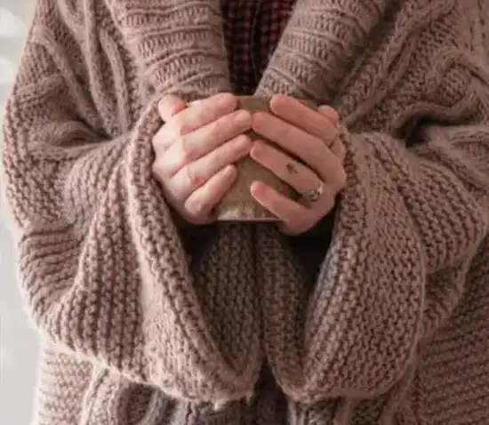 Cozy oversized taupe chunky knit cardigan worn with a red plaid shirt underneath, hands wrapped around a warm mug
