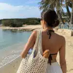 Behind view of woman wearing black swimsuit top with white linen shorts holding a crochet market bag standing on a sandy beach with palm trees in the background.