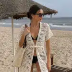 Woman wearing a crochet tie front-beach cover up over a black bikini, wearing black shades, holding a market bag and stand on a sandy beach.