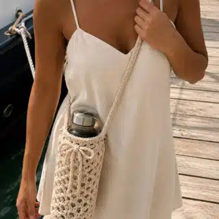 Woman wearing a cream color summer slip dress with a Crochet Water Bottle Crossbody Holder standing on a dock with a sail boat in the background.