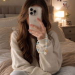 Woman wearing a sweatshirt and sweatpants taking a selfie in a bedroom with a coastal charm beaded wristlet attached to the smartphone.