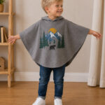 Young boy wearing a gray hooded poncho made from an upcycled graphic T-shirt featuring a mountain and forest print. The relaxed poncho is paired with blue jeans and white sneakers, creating a cozy and beginner-friendly DIY sewing project for kids.