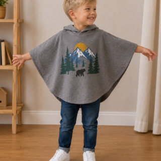 Young boy wearing a gray hooded poncho made from an upcycled graphic T-shirt featuring a mountain and forest print. The relaxed poncho is paired with blue jeans and white sneakers, creating a cozy and beginner-friendly DIY sewing project for kids.