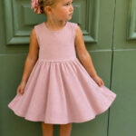 Young girl wearing a blush pink sleeveless twirly play dress made from an upcycled oversized T-shirt. She stands in front of a sage green door wearing white sneakers, holding the full gathered skirt to show its playful twirl and comfortable fit.