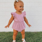 Toddler girl wearing a soft pink ruffle-sleeve romper made from an upcycled T-shirt. The knit romper features flutter sleeves, an elastic waist, snap crotch closure, and white sandals, making it a comfortable outfit for everyday play.