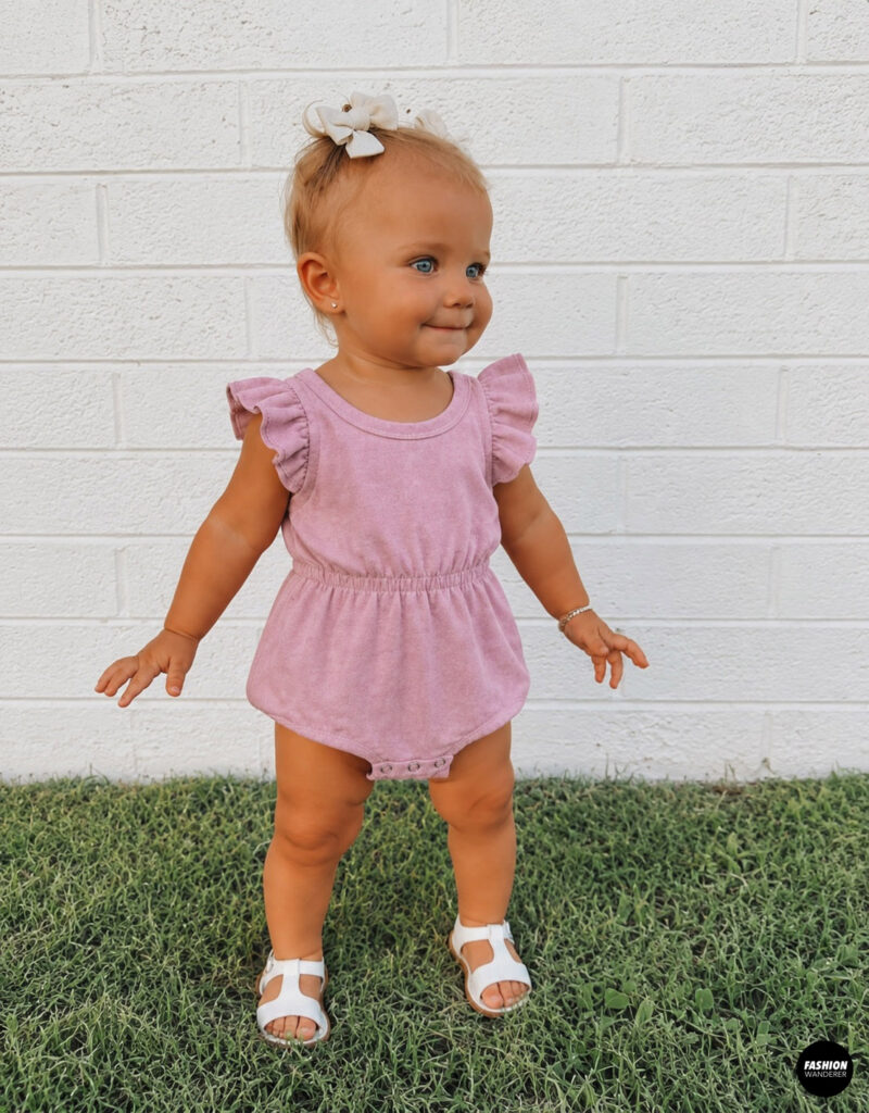 Toddler girl wearing a soft pink ruffle-sleeve romper made from an upcycled T-shirt. The knit romper features flutter sleeves, an elastic waist, snap crotch closure, and white sandals, making it a comfortable outfit for everyday play.