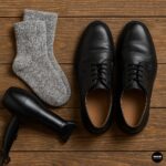 Gray wool socks, black leather dress shoes, and a black hair dryer on a wooden surface for shoe stretching preparation.