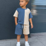 Toddler boy modeling a two-tone blue and cream color-block T-shirt and matching shorts set sewn from recycled T-shirts. The casual outfit features a relaxed fit, elastic drawstring shorts, and white sneakers for a modern everyday look.
