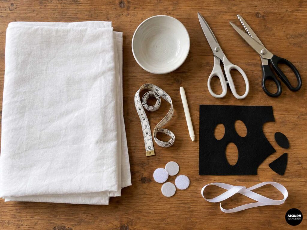 Flat lay of materials for a DIY no-sew dog ghost costume, including white fabric, measuring tape, fabric scissors, pinking shears, black felt, white ribbon, hook-and-loop dots, chalk, and a small bowl.