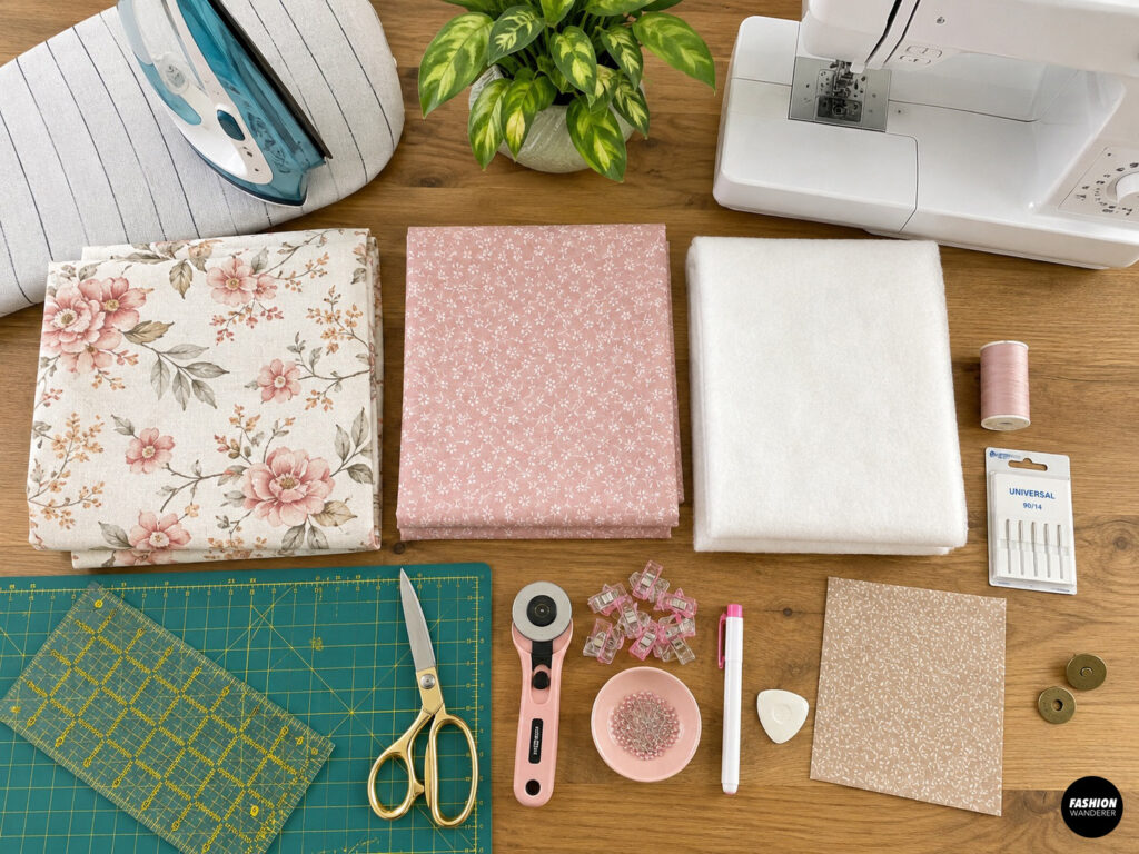 Flat lay of materials for making a DIY quilted bow tote bag, including floral exterior and lining fabrics, batting, sewing machine, iron, thread, needles, ruler, rotary cutter, scissors, clips, pins, marking tools, and magnetic snaps.