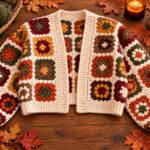 Fall granny square cropped cardigan crochet pattern in cream, rust, mustard, burgundy, olive, and brown yarn displayed on a wooden table with autumn leaves, yarn, pumpkin, candle, and coffee.
