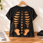 Finished black T-shirt transformed into a skeleton rib cage cutout top hanging on a wooden hanger with pumpkins, plants, scissors, and a paper skeleton template nearby.