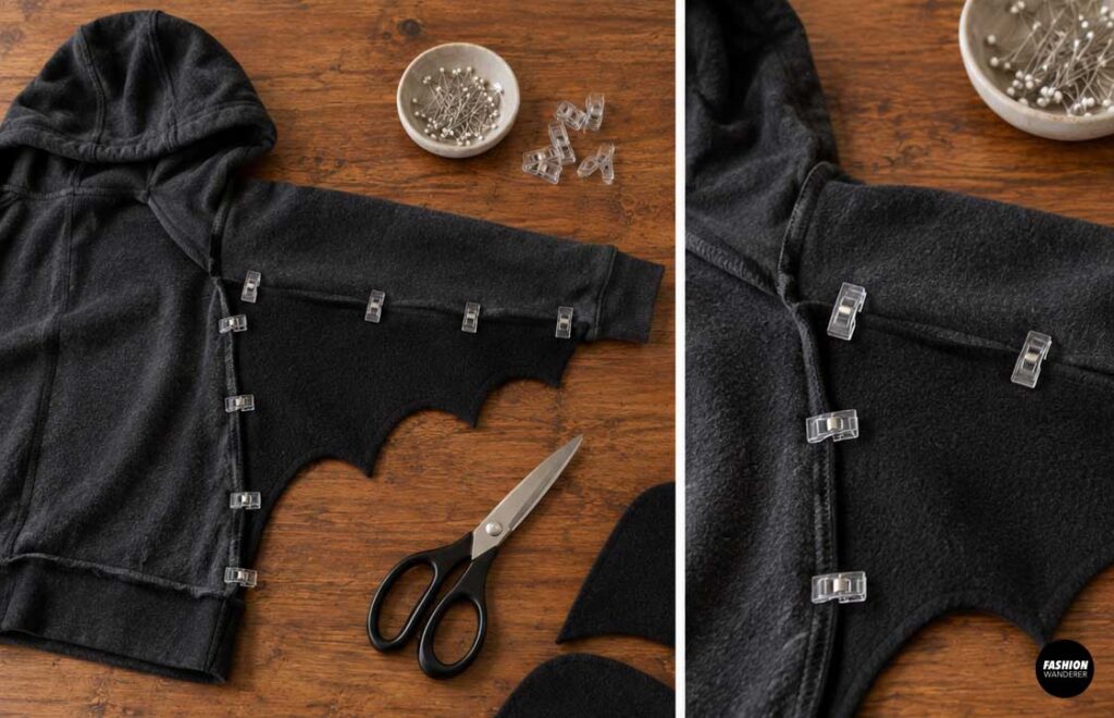 Black fabric bat wing clipped between the hoodie body and sleeve, demonstrating how to position the wing before sewing.