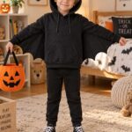 Young boy wearing a black hoodie and matching pants transformed into a bat costume with pointed ears and fabric wings attached under the sleeves, holding an orange jack-o&rsquo;-lantern trick-or-treat bag in a Halloween-decorated kids&rsquo; bedroom.